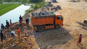 Мини-бульдозер продвигает камни и валуны в воду, а самосвал выгружает грунт.