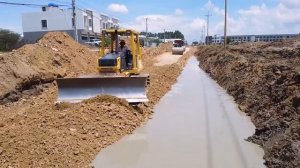 Грузовики и бульдозер засыпают канал вдоль дороги.