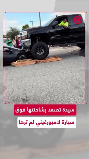 سيدة تقود شاحنة ضخمة وتصعد فوق سيارة لامبورغيني فاخرة في موقف سيارات وصاحبها داخلها.