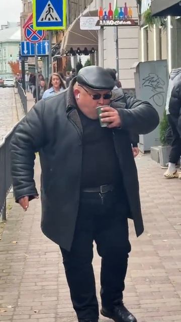 Полный мужчина в черной кожаной куртке в очках идет по улице со стаканом кофе в руке  мем мемы